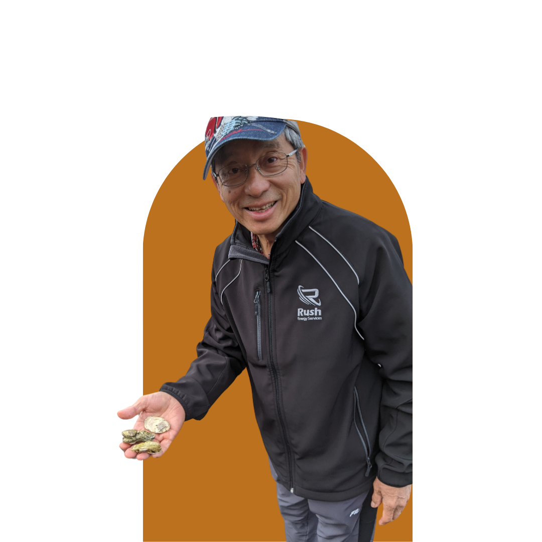A smiling man wearing glasses and a black Rush Energy Services jacket, holding a handful of small seashells or rocks, standing against a brown background.
