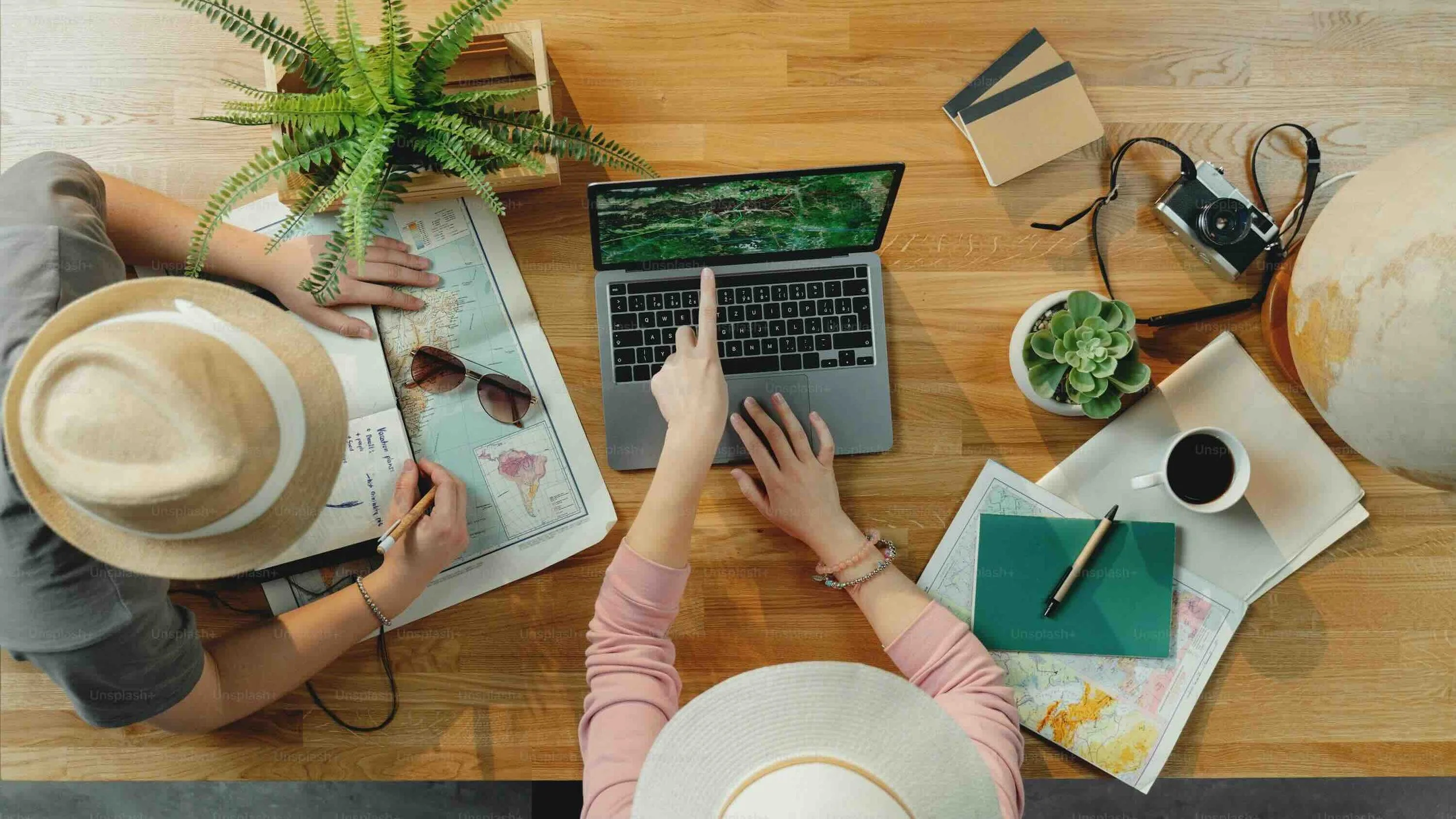 Top-down view of a wooden table with two women working on a travel planning project. One woman, wearing a hat, is pointing at a map. The other is touching a laptop with a forest background on the screen. The table has travel books, a camera, a globe, a coffee cup, a small potted plant, sunglasses, and notebooks.