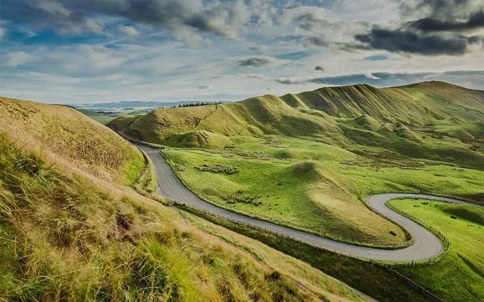 A winding road cutting through lush green rolling hills under a partly cloudy sky.