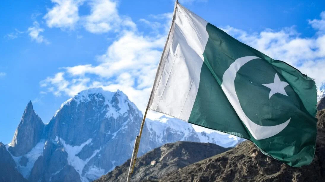 Pakistani flag flying against snow-capped mountains and a blue sky with clouds.