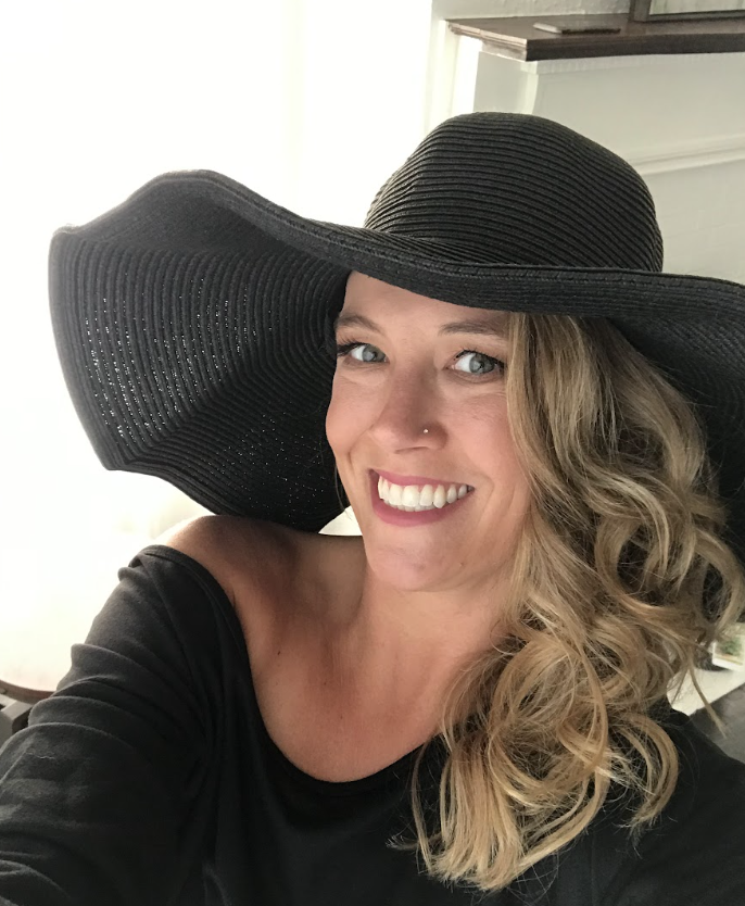 A woman with blonde curly hair, smiling, wearing a large black sun hat and black top.