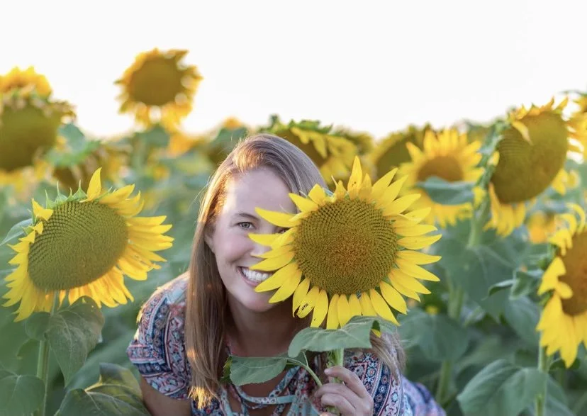 A woman smiling in a sunflower field holding a sunflower close to her face.