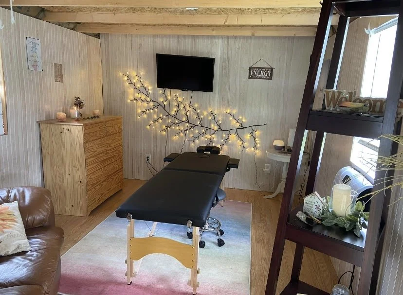 A massage therapy room with a massage table, a wall-mounted TV, string lights shaped like tree branches, a wooden dresser, a wicker chair, and a dark bookshelf with plants and decorative items. The room has light-colored wood-paneled walls and ceilin