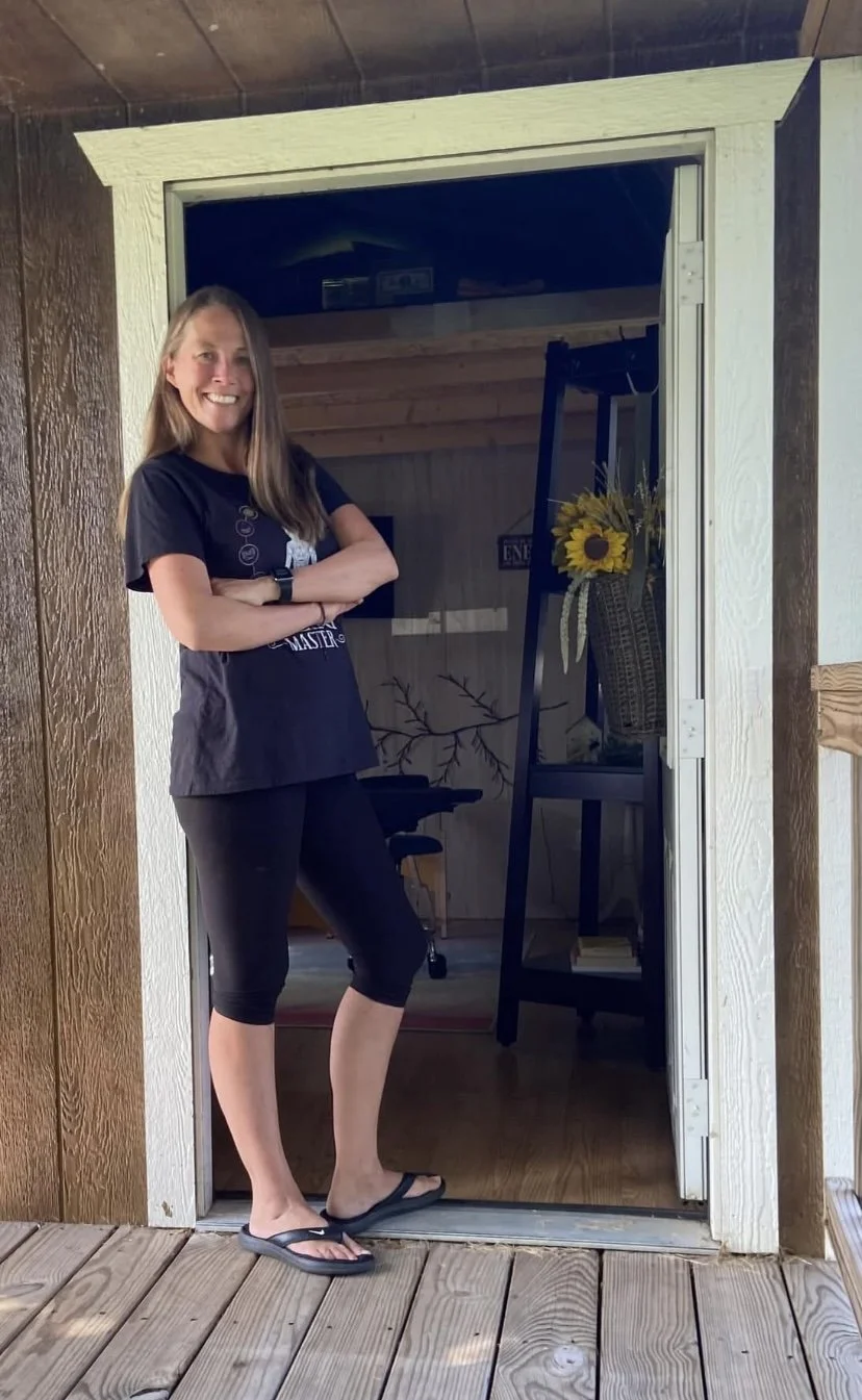 Woman standing at a doorframe, smiling, wearing black leggings, a black T-shirt, and flip-flops, with a porch and interior room visible behind her.
