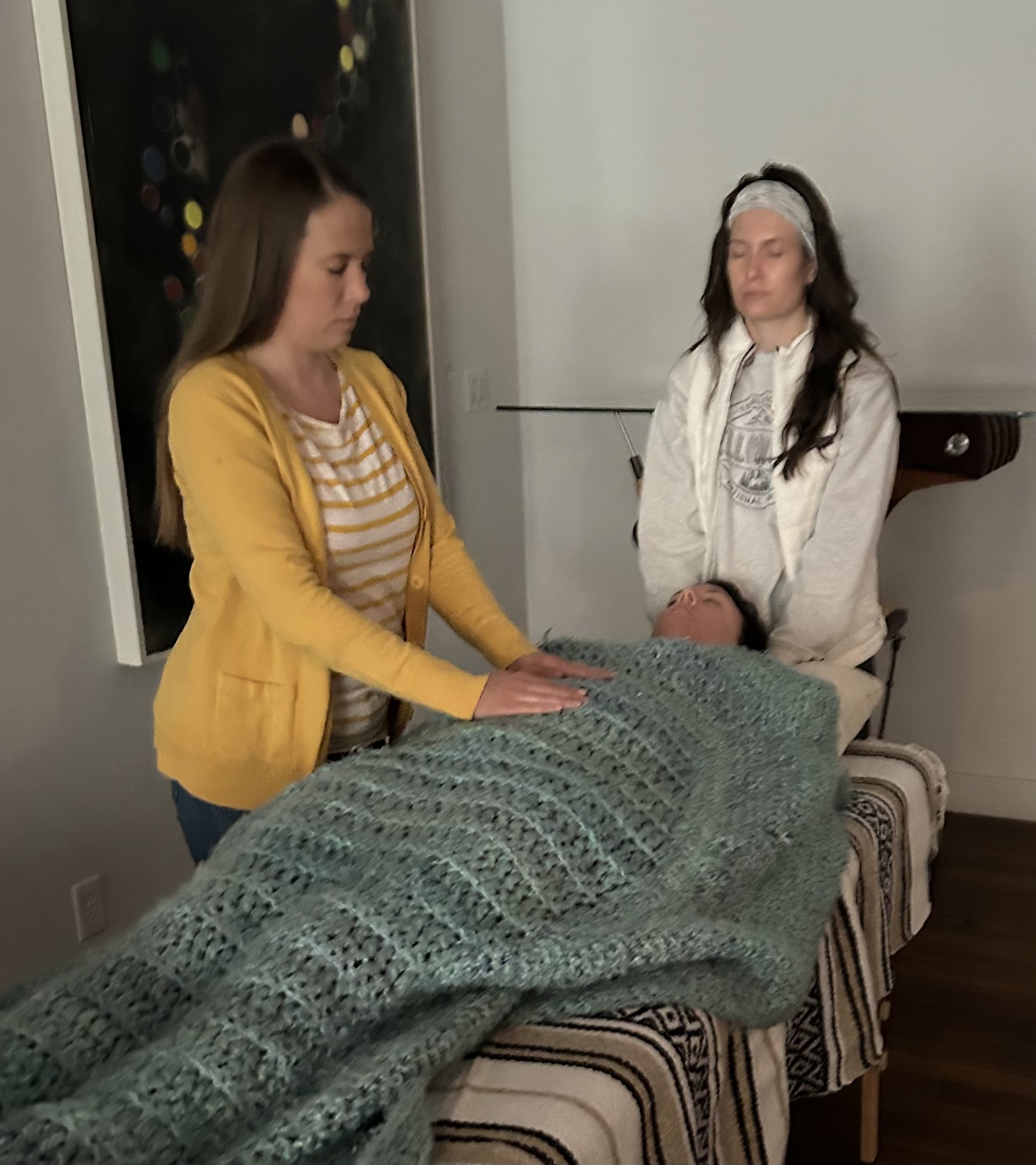 Two women standing by a person lying on a table, with one woman placing her hands on the person's back. The person on the table has dark hair and their face is partially visible. The woman on the left is wearing a yellow cardigan and striped shirt, while the woman on the right is wearing a light-colored headband and sweatshirt.