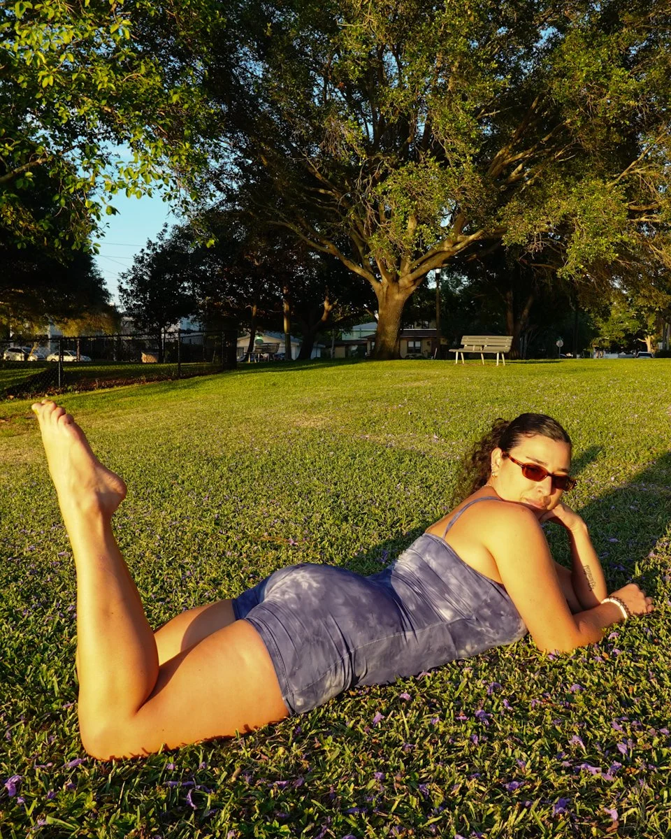 A woman lying on grass in a park, wearing sunglasses and a blue dress, with a large tree in the background during sunset.