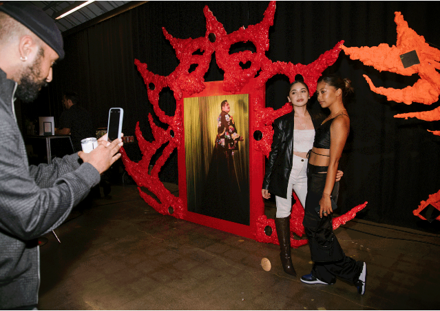 Two women posing for a photo at a Halloween event with a red and black spooky backdrop. A man is taking their picture with a smartphone.
