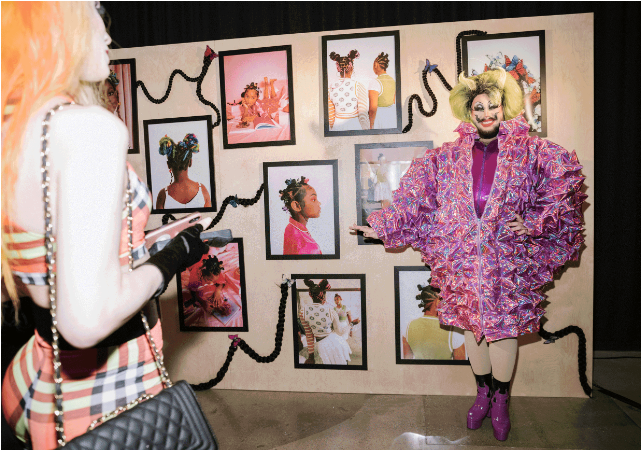 Colorful clown with yellow hair, purple ruffled jacket, and purple boots posing in front of a wall decorated with framed photographs of children with styled hair.