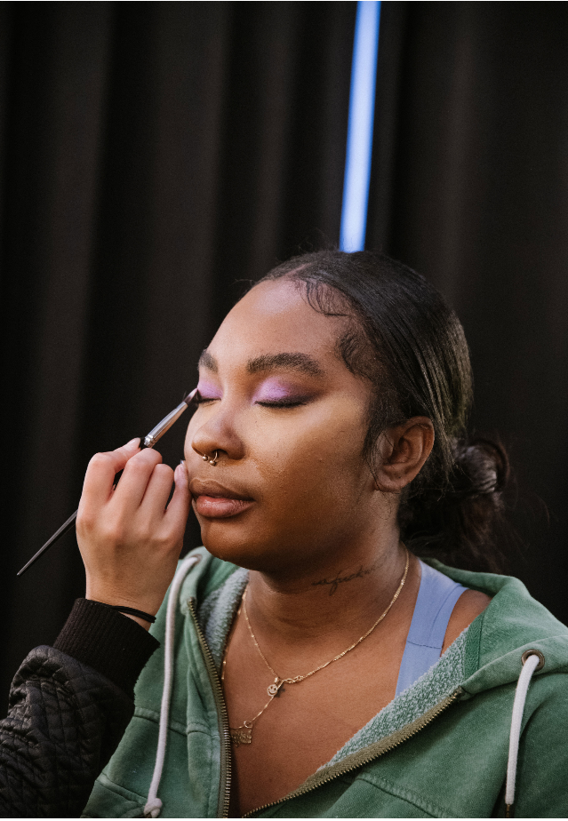 A woman with closed eyes is having makeup applied with a brush by another person. She wears a green hoodie, a necklace, and has purple eyeshadow.