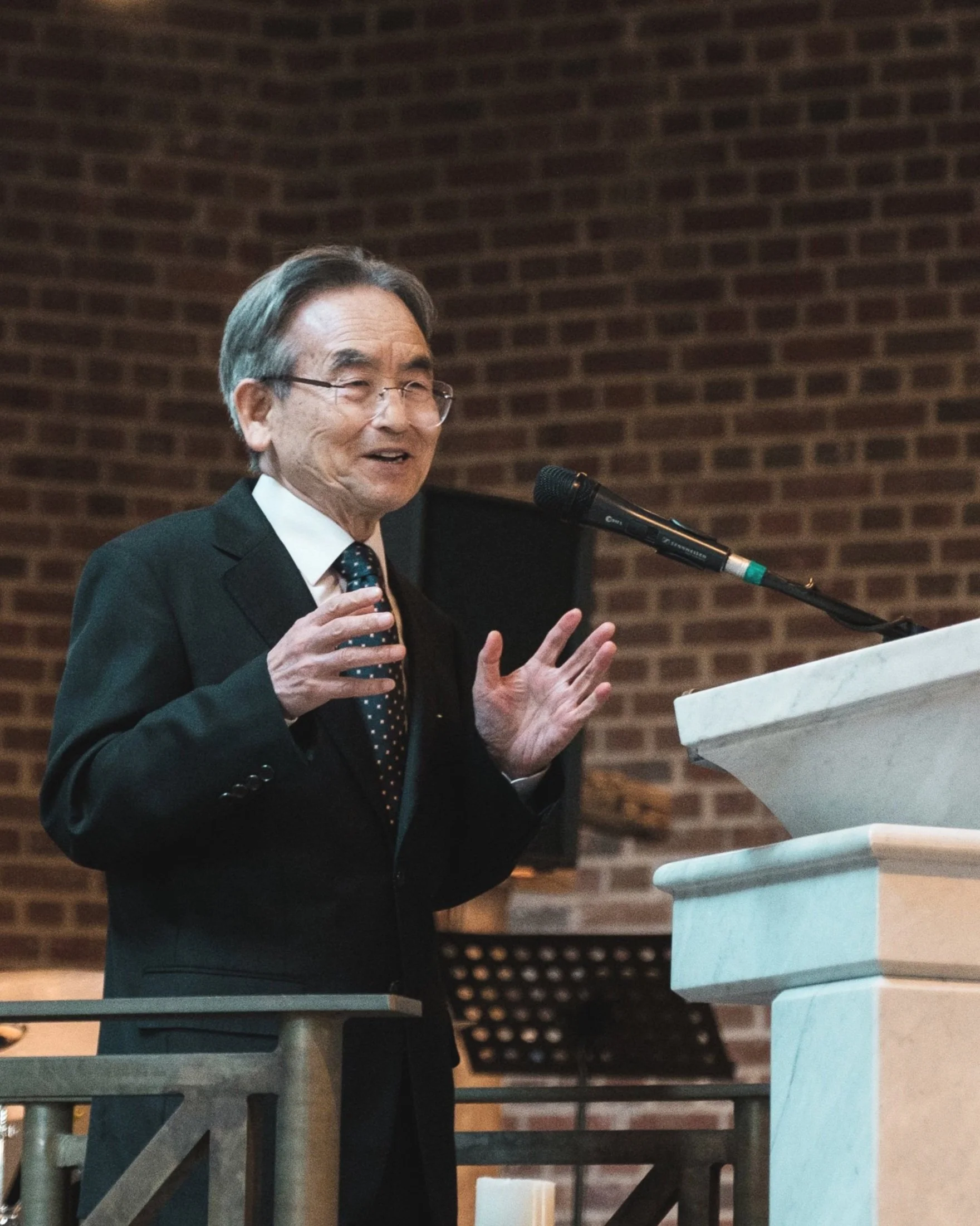 Ein älterer Mann in Anzug hält eine Rede an einem Rednerpult in einer Kirche mit Backsteinmauer im Hintergrund.