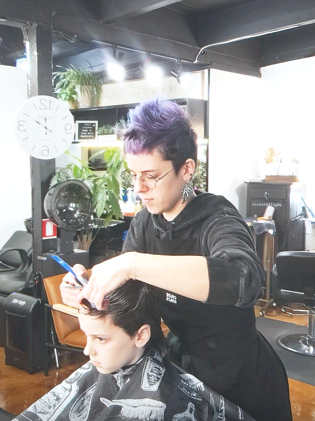 A hairstylist cutting a young man's hair in a salon. The hairstylist has short purple and black hair and is wearing glasses and earrings. The young man has dark hair and is sitting in a barber chair with a cape. The salon has plants, a clock, and various salon equipment visible in the background.