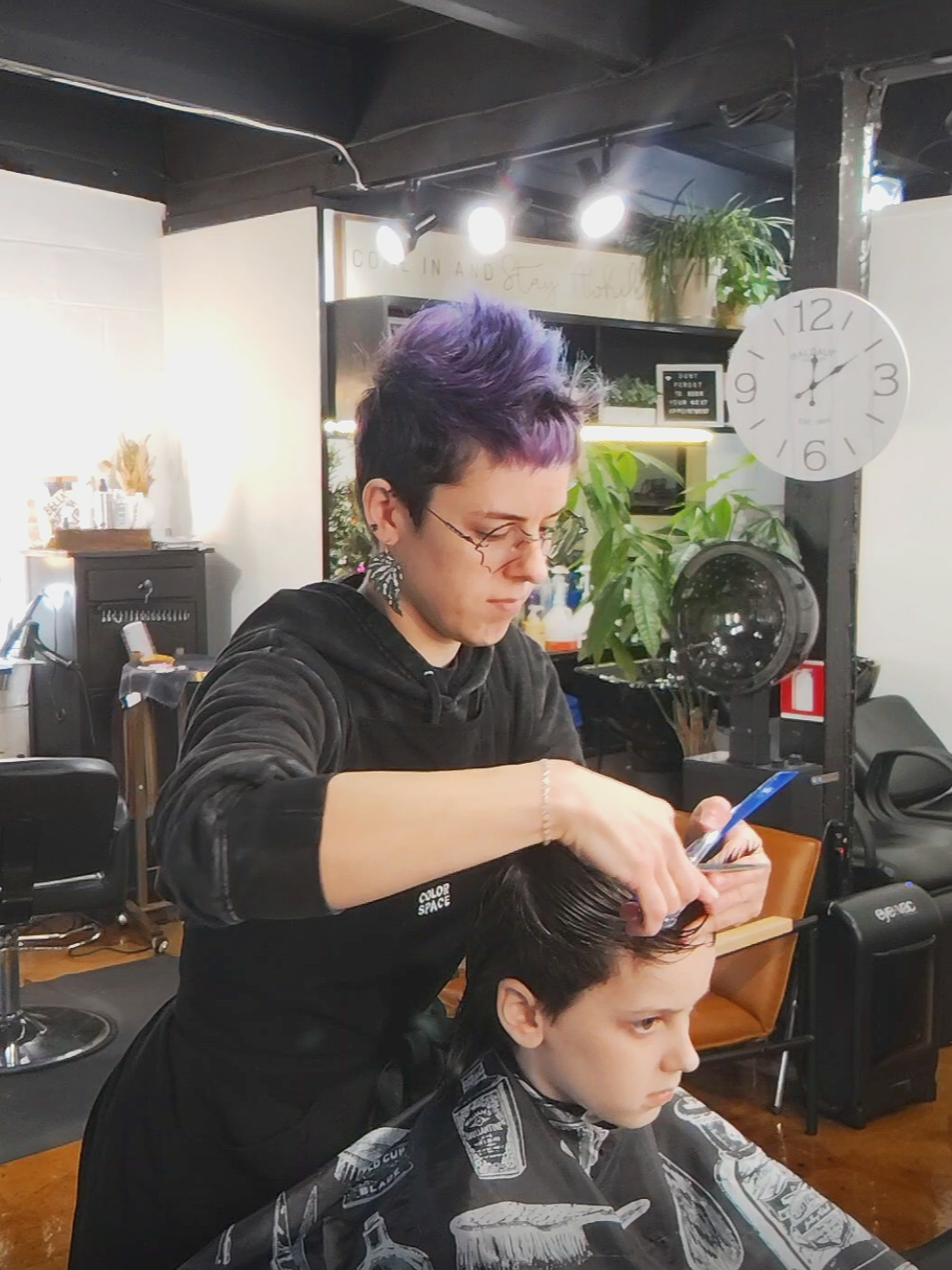 Barber cutting a young boy's hair in a salon, with plants and a clock on the wall in the background.