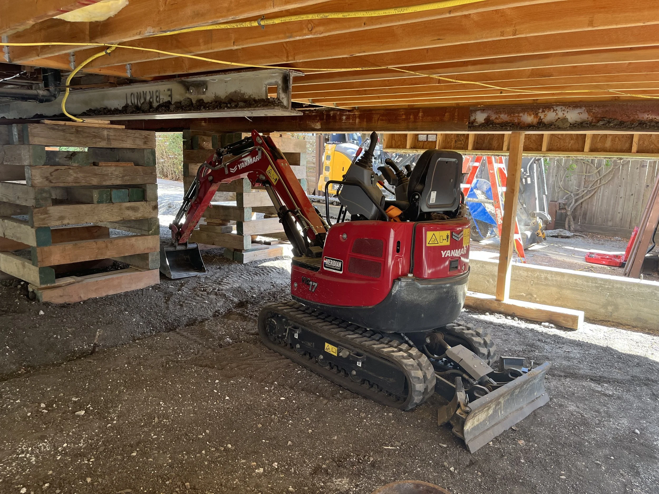 A house lifted into the air, supported by I beams and wooden cribbage. A mini excavator is underneath the lifted structure, so workers can dig new footings underneath the house to replace the foundation.