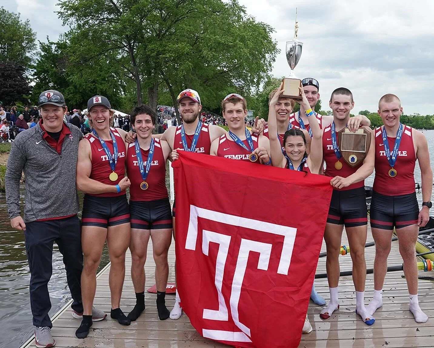 A group of nine rowers and a coach celebrating with medals and trophies on a dock, holding a red and white flag with a large stylized T, after a rowing competition.