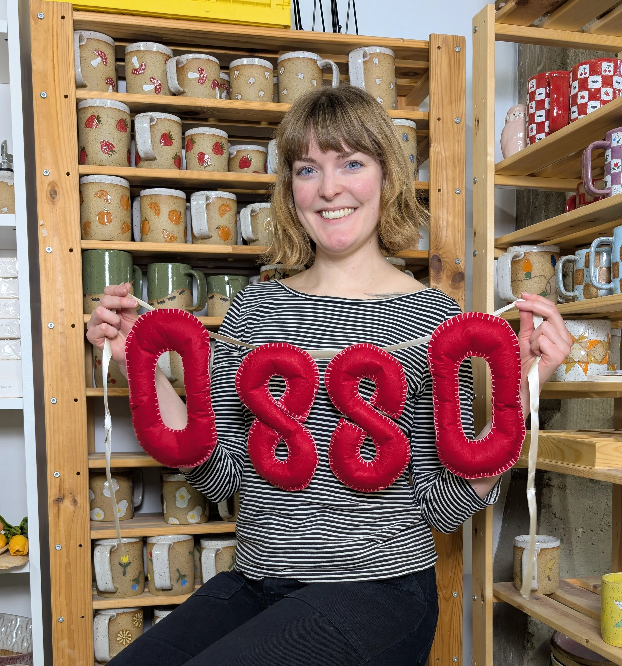 Person holding a banner that says OSSO in red letters. She is in front of shelves with finished pottery..