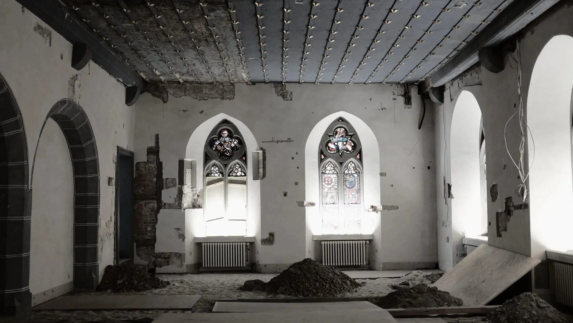 Interior of a church under renovation with exposed bricks, torn walls, and construction dirt on the floor. Two stained glass windows are visible on the far wall, with some sections of the wall missing or damaged.