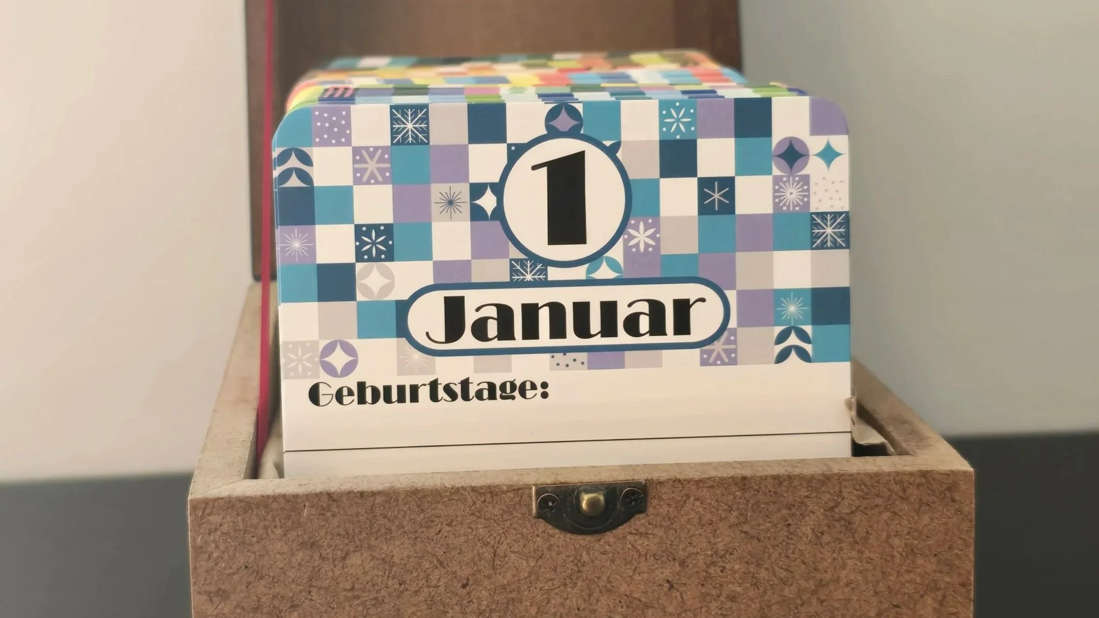 Mit Postkarten gefüllte Holzkiste mit unterem Teil eines Kofferschlosses, die sichtbare Karte zeigt den 1. Januar, das Hintergrundmuster für diesen Monat zeigt eine Mischung aus blau-weißen Kästchen, teilweise mit Schneeflocken und Eiskristallen verz
