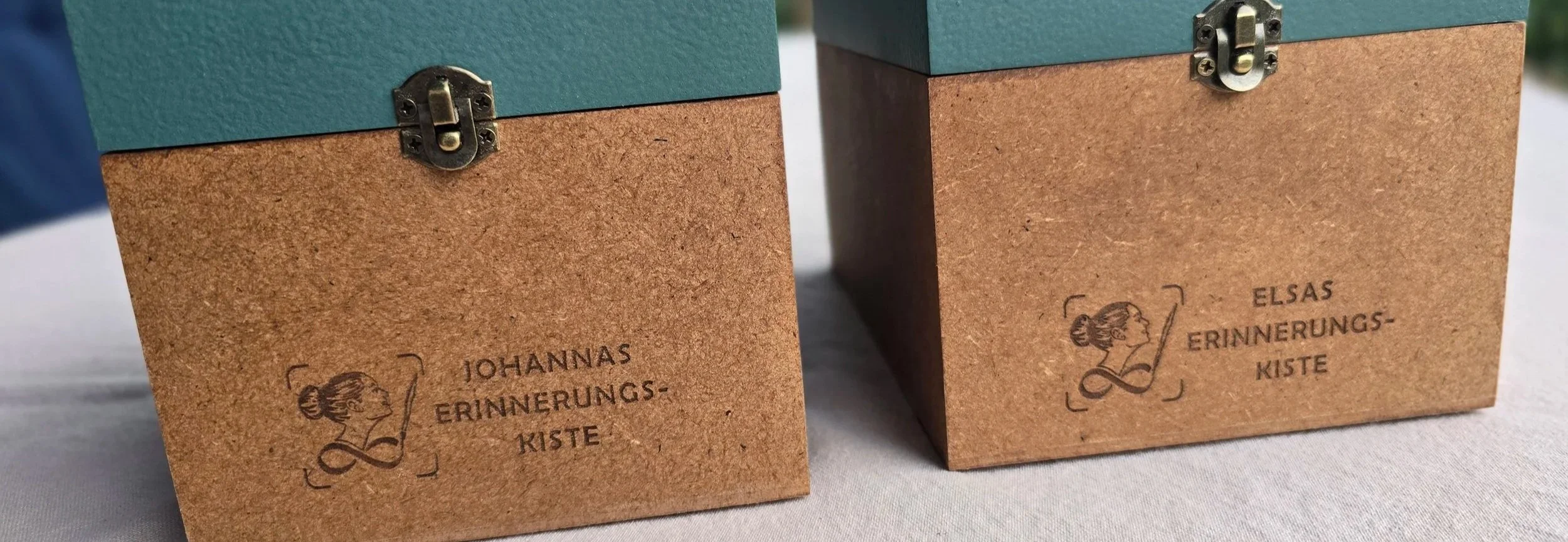 Auf dem Foto sind die Unterteile zweier Kisten mit den Gravuren für die Erinnerungskisten zu erkennen, es sind neben dem Logo der Erinnerungskisten die Namen Elsa und Johanna in das Holz graviert. Die Gravur hebt sich dunkel von dem MDF-Holz ab und i