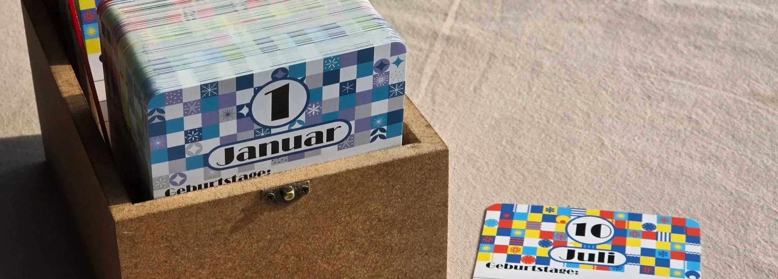 Das Bild zeigt auf der linken Seite eine geöffnete Erinnerungskiste, welche mit den Postkarten gefüllt ist, sichtbar ist der 1. Januar. Neben der Kiste liegt die Karte des 10 Juli. Der Januar hat im Hintergrund eine blau-weiße Musterung, der Juli ist
