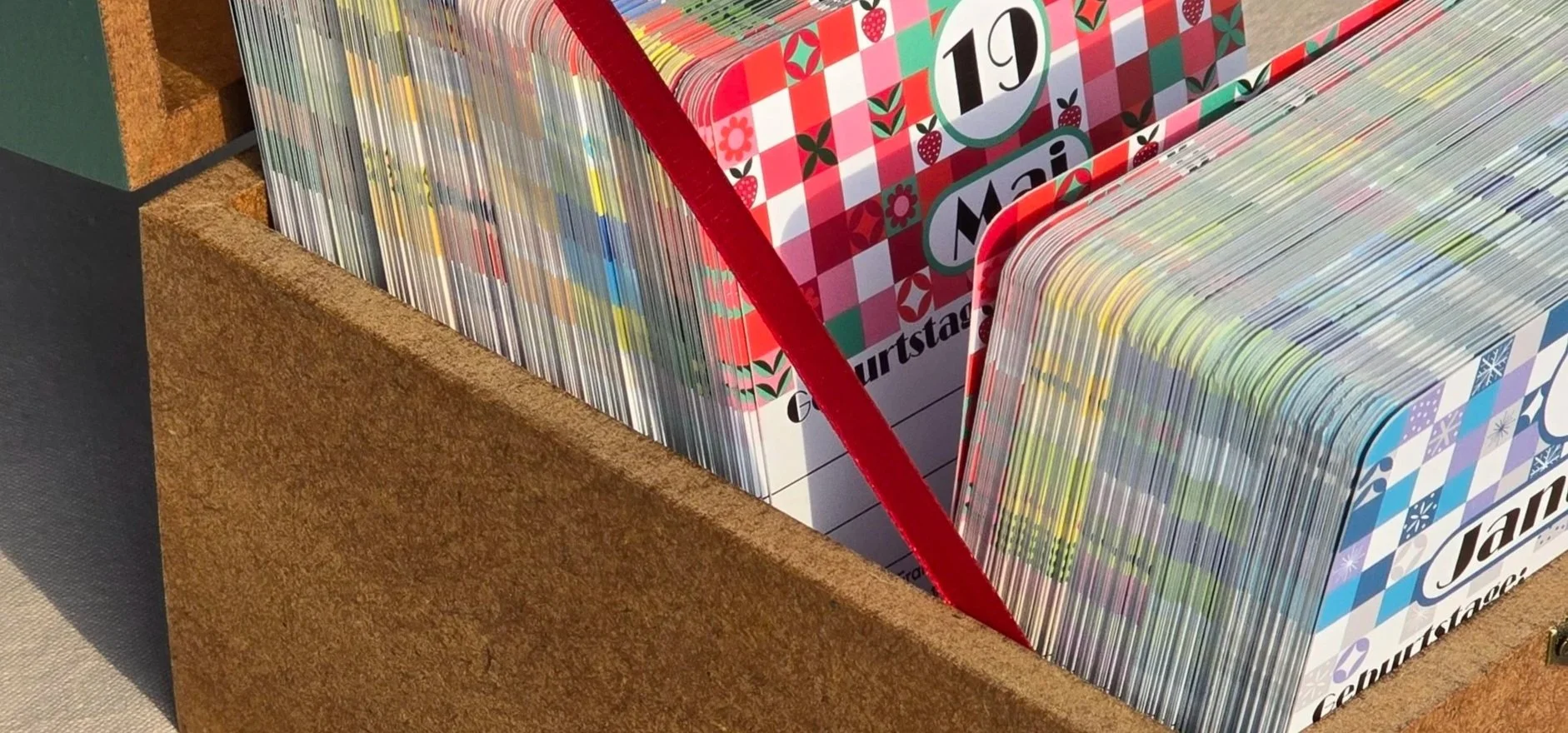 Das Foto zeigt das Unterteil einer Kiste aus MDF-Holz, welches mit Postkarten gefüllt ist. Sichtbar sind die Postkarten des 19. Mai mit einer rot-weißen Musterung und Verzierungen durch stilisierte Blumen, Blätter und Erdbeeren und der 1. Januar mit 
