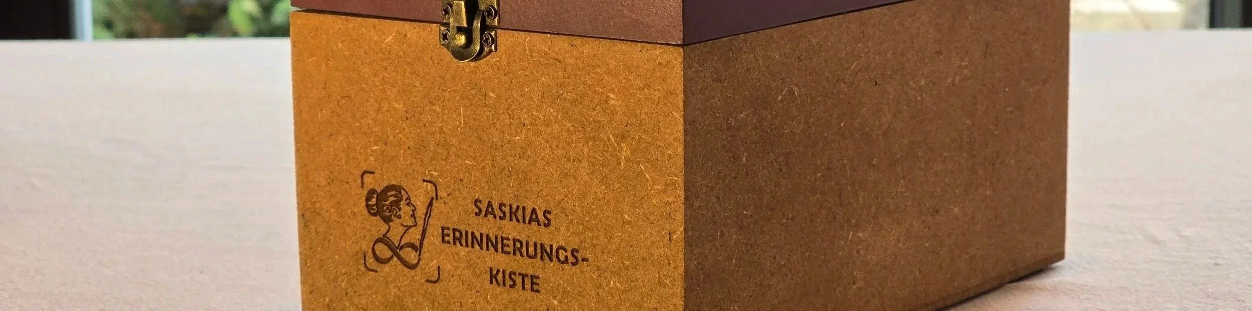 Eine braune Holztruhe mit einem goldfarbenen Schloss auf einem hellen Tisch, mit der Aufschrift 'Saskias Erinnerungs-Kiste' und einer Illustration einer lachenden Frau.
