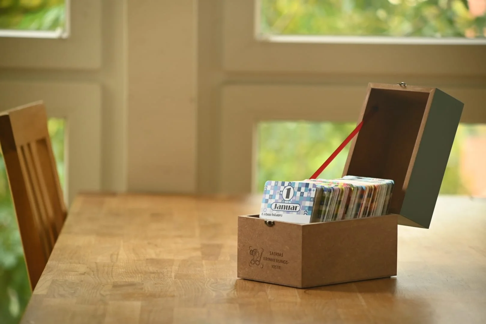 Holzkiste mit rotem Band, geöffnet auf einem Tisch vor einem Fenster mit Blick ins Grüne. Sichtbar ist die Karte für den 1. Januar. Fronseitig ist die gelaserte Aufschrift „SASKIAS ERINNERUNGS-KISTE“ sichtbar.