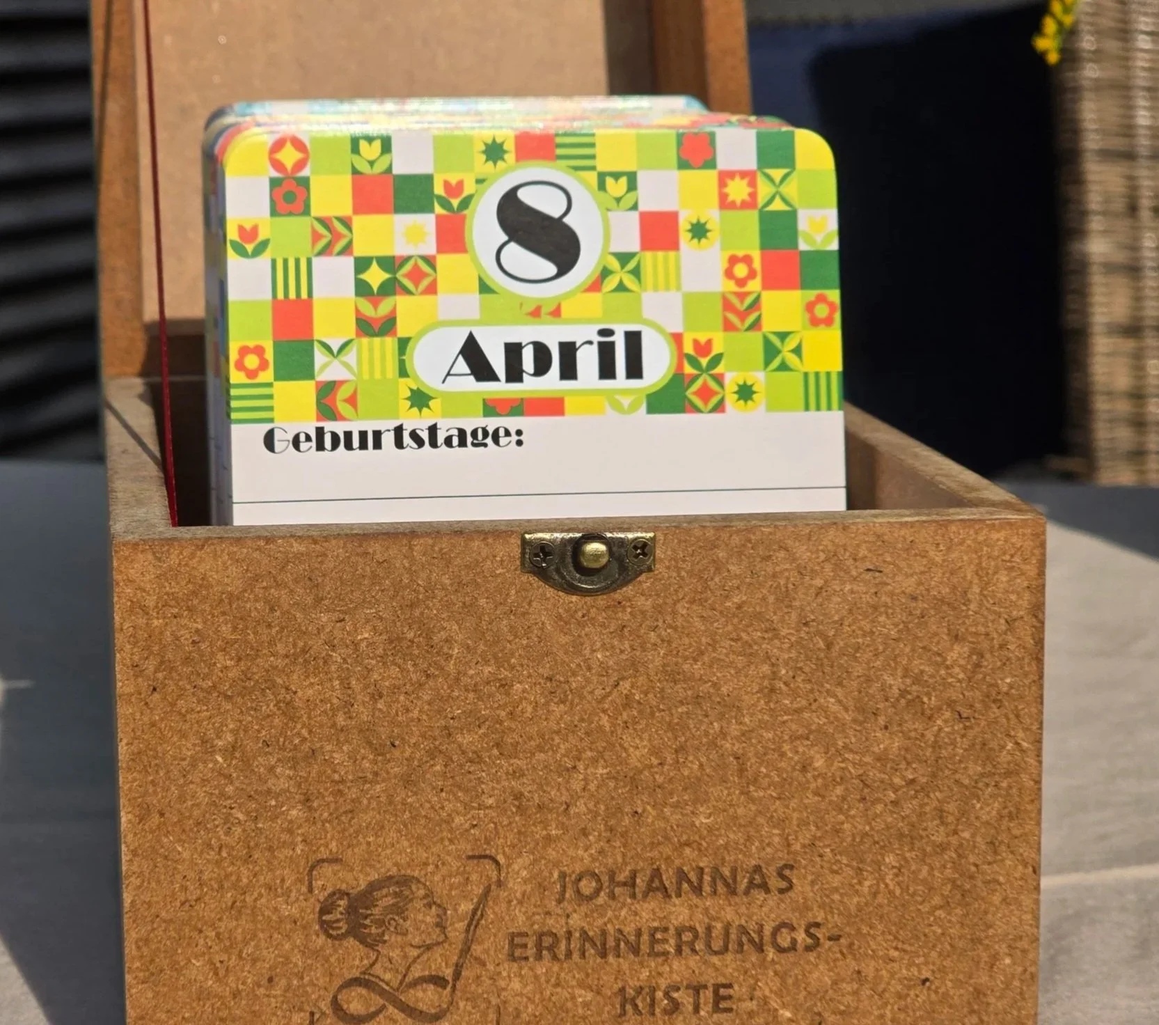 Eine geöffnete Erinnerungskiste aus Holz steht auf einem Tisch, sichtbar ist die gelb-grün gemusterte Kalenderkarte des 8. April. Die Kiste ist graviert mit dem Namen Johanna.