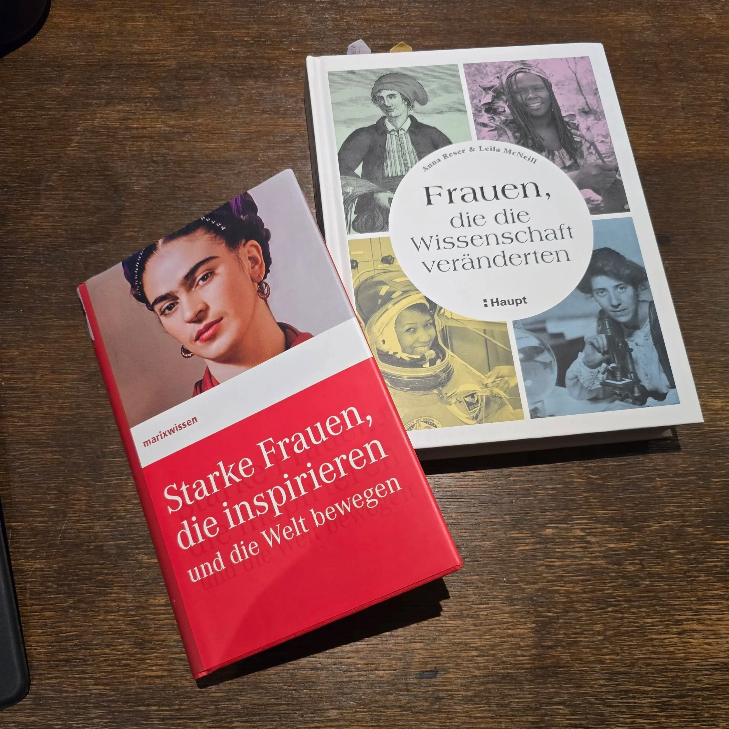 Zwei Bücher liegen auf einem dunklen Holztisch. Das erste Buch handelt von inspirierenden Frauen der Weltgeschichte, das andere von Frauen, die wichtige Errungenschaften in der Wissenschaft erlangten.