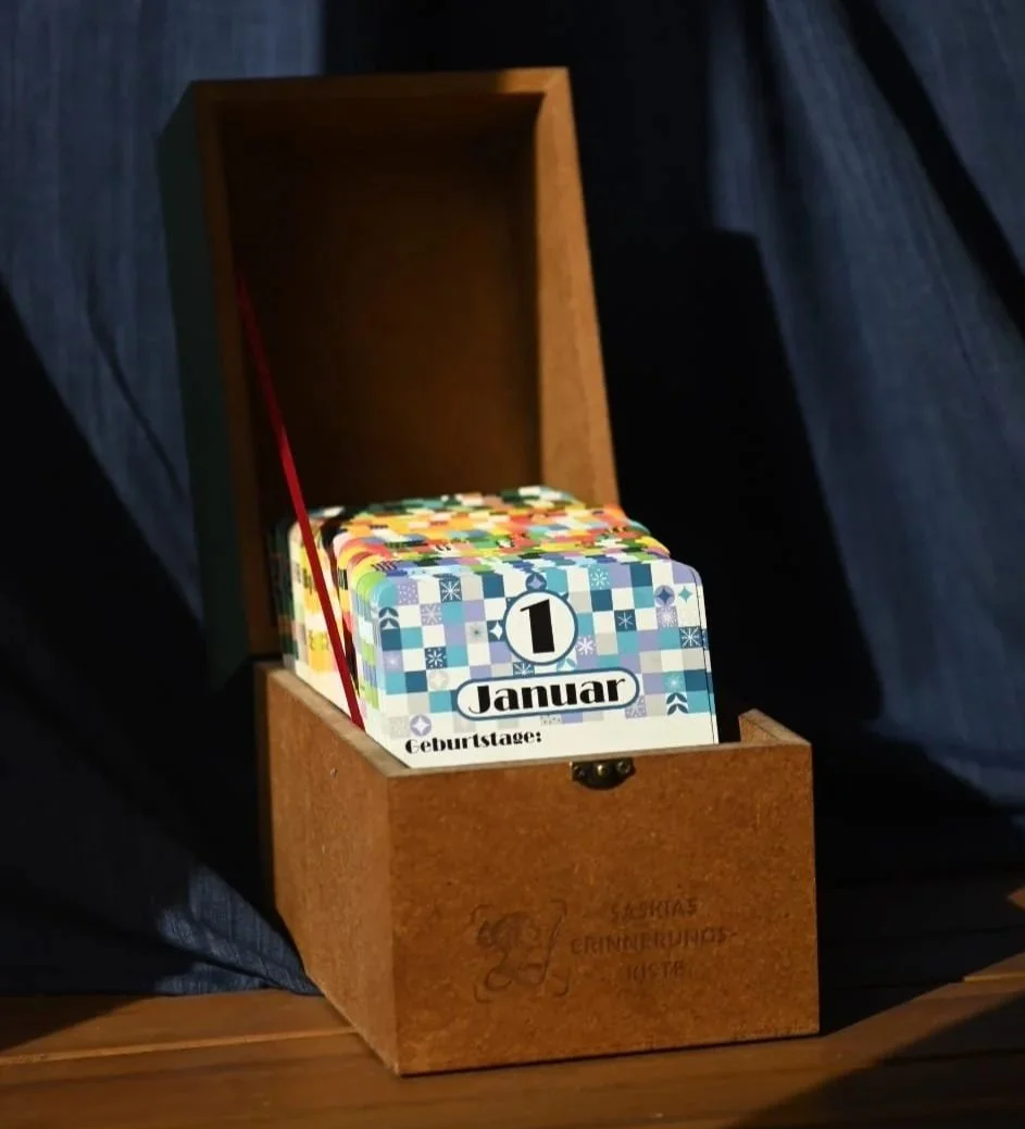 Geöffnete Erinnerungskiste auf einem Holztisch stehend vor dunklem Hintergrund. Sichtbar ist die blau-weiß-gemusterte Karte vom 1. Januar.