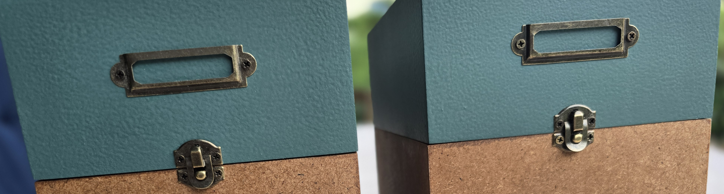 Zwei blaue Aufbewahrungsboxen mit Metallgriffen und Schloss, eine steht auf einem braunen Karton, im Hintergrund ist unscharf Grün zu erkennen.