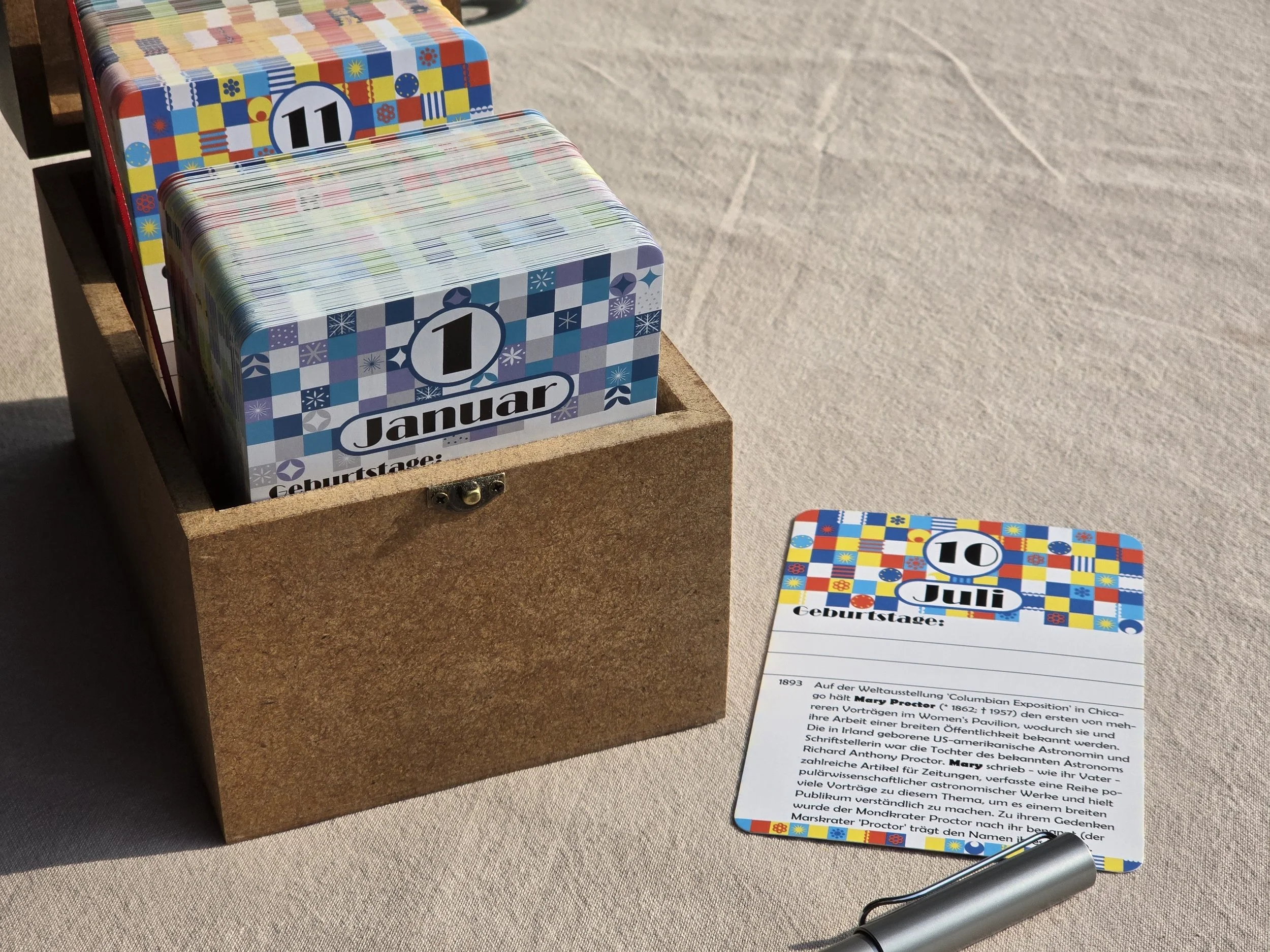 Kalenderkarten für den 1. Januar und den 10. Juli, mit buntem Muster, in einer Holzkiste, auf einer Tischdecke liegend, neben einem Metallstift und einer Textkarte.