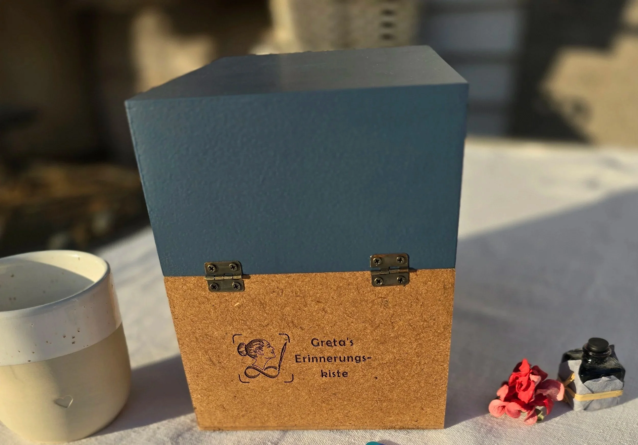 Eine Erinnerungskiste aus Holz mit blaugrauem Deckel und Scharnieren, mit der Aufschrift ‚Greta's Erinnerungskiste‘. Neben der Kiste befinden sich eine weiße Tasse, rote Blumen und eine kleine Flasche auf einer weißen Tischdecke.