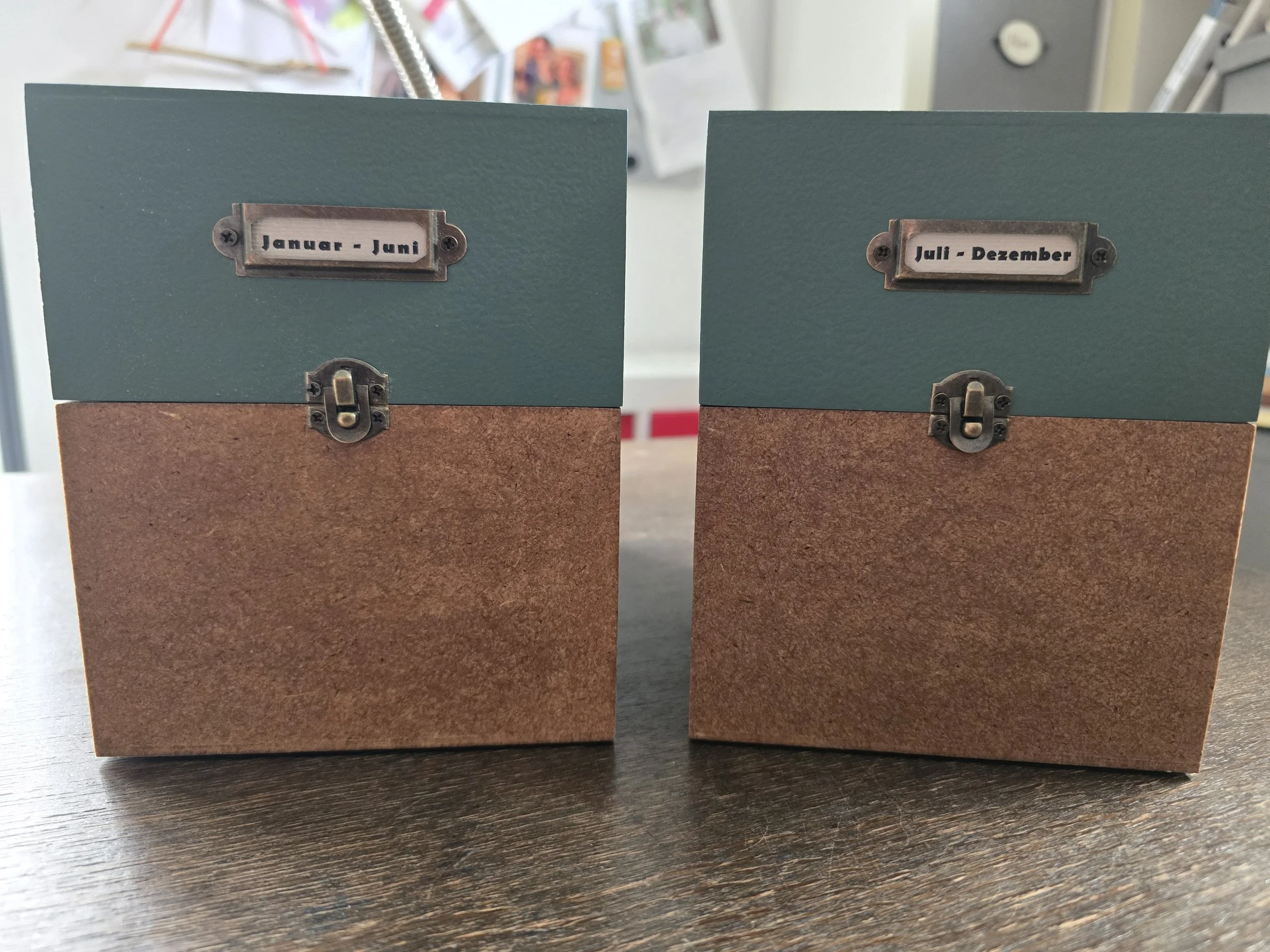Zwei handgefertigte Holzkisten mit Metallverschluss und Monatsbeschriftung „Januar – Juni“ sowie „Juli – Dezember“ stehen nebeneinander auf einem Schreibtisch. Die Kisten dienen zur Aufbewahrung der Kalenderkarten und weiterer Erinnerungsstücke.