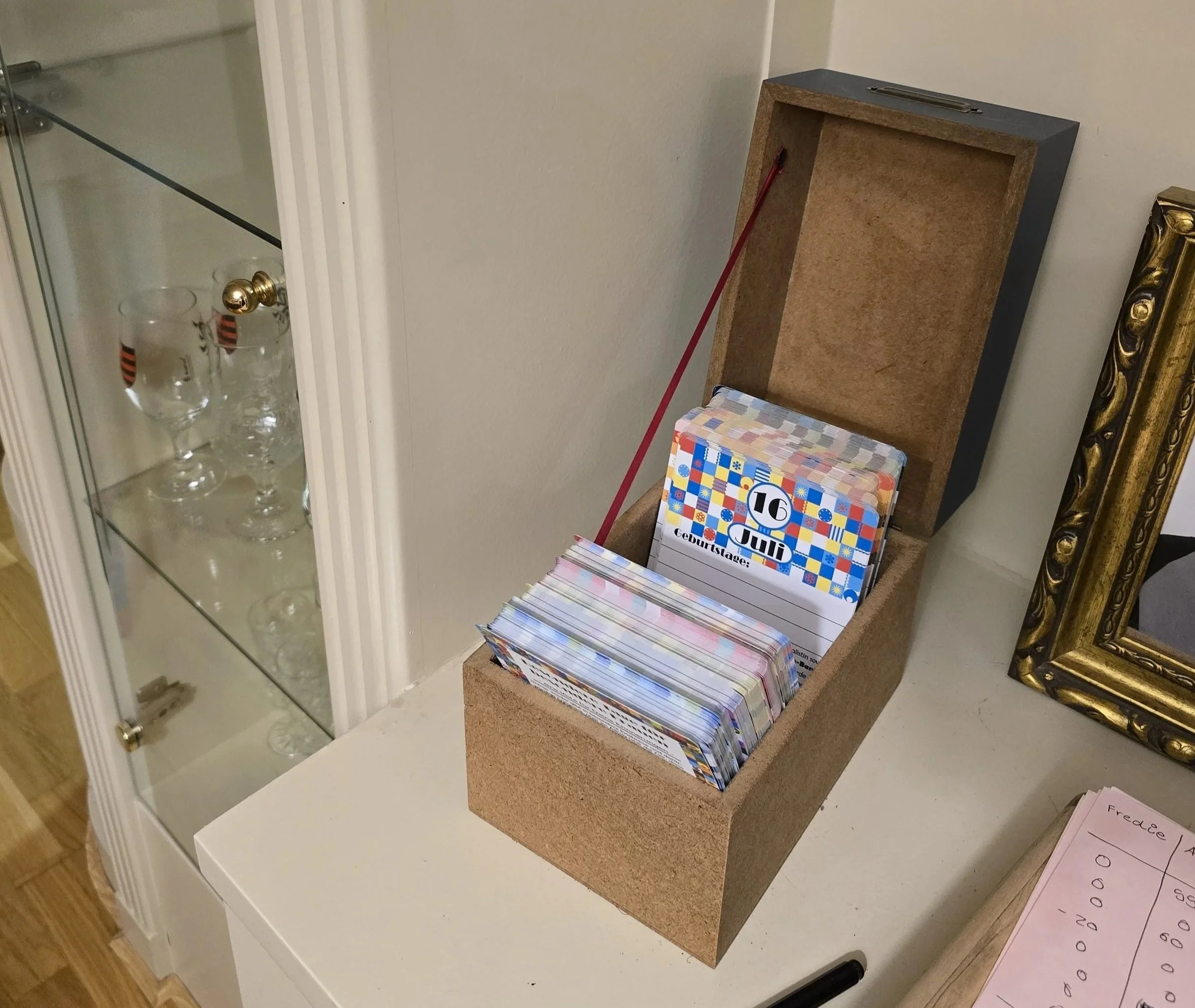 Ein offenes Kartenbox mit Geburtstagskarten und anderen Karten ist auf einem Tisch. Im Hintergrund sind Gläser in einer Vitrine zu sehen.