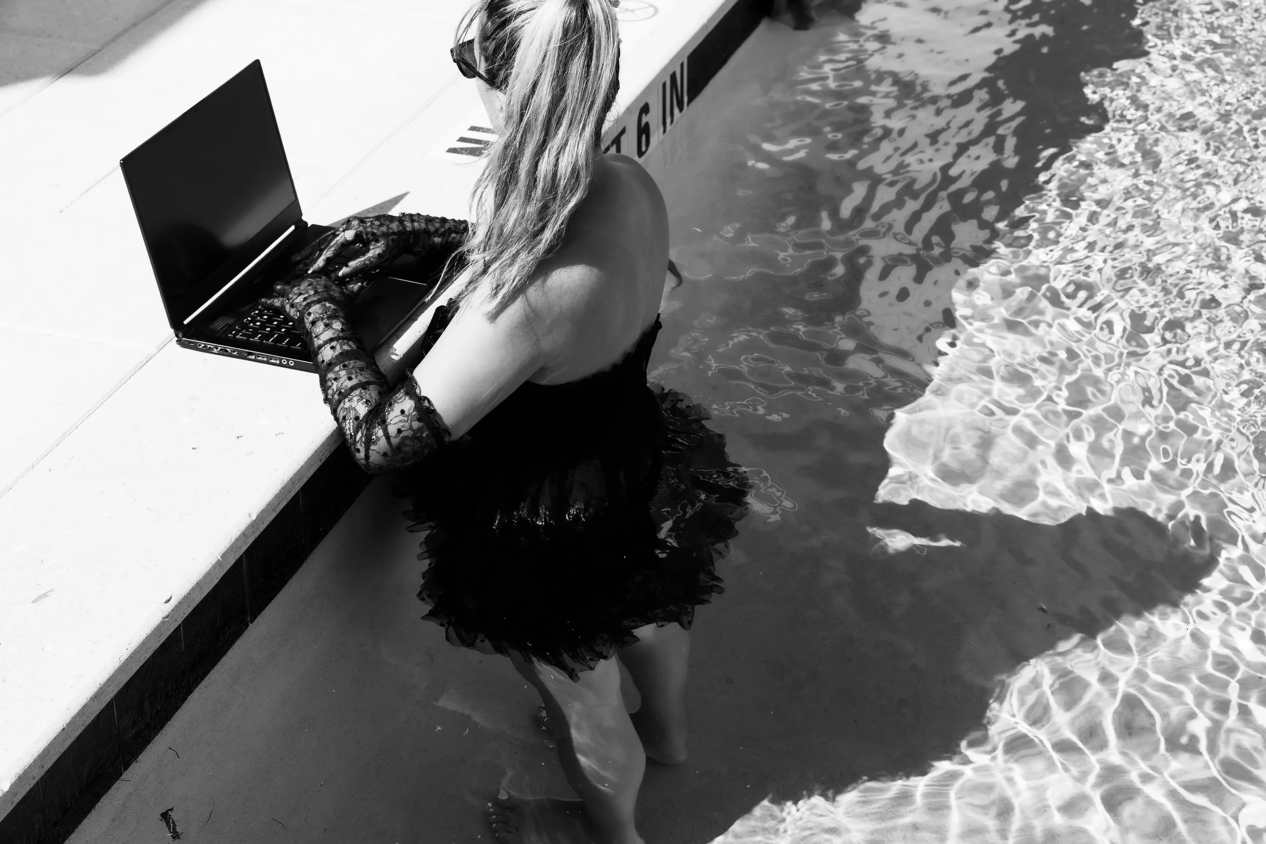 A woman in a black dress stands waist deep in a pool while she writes a blog for SEO on a laptop on the edge