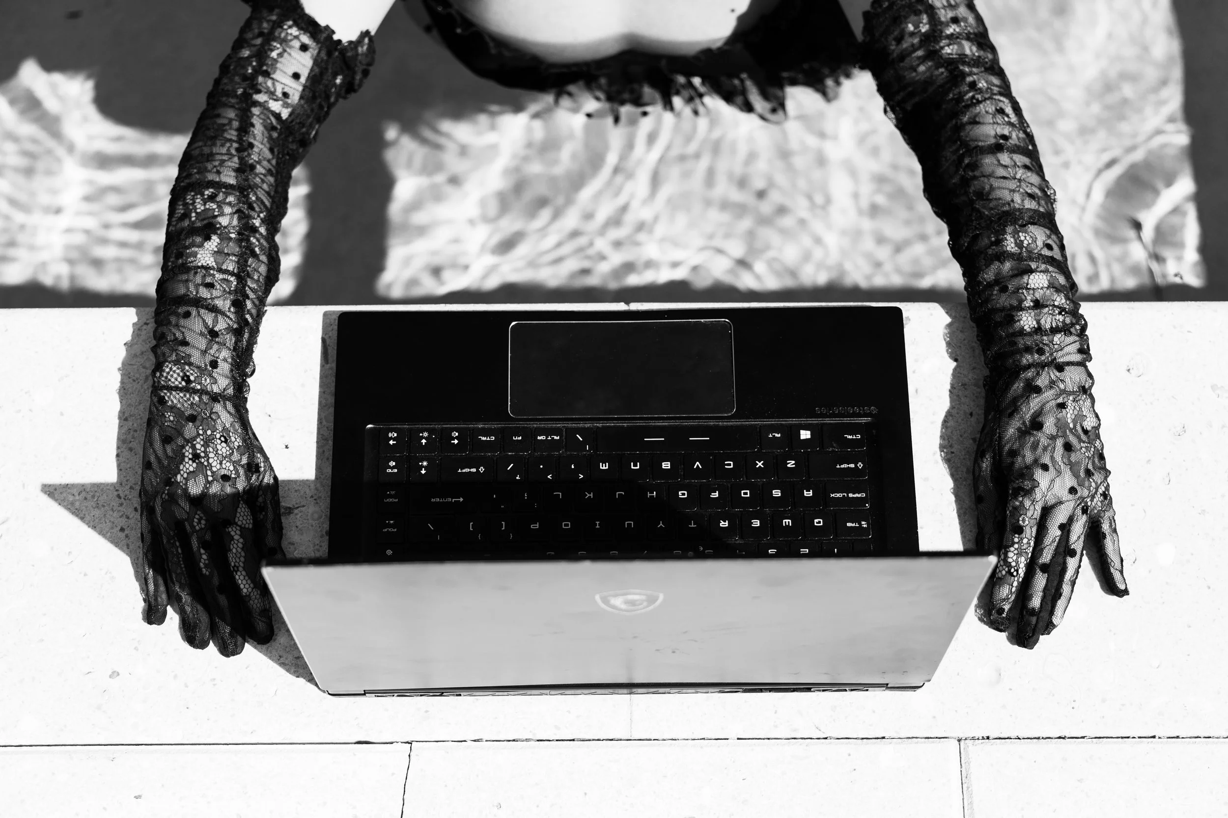 A woman with lace gloves stands by a laptop on the edge of a pool