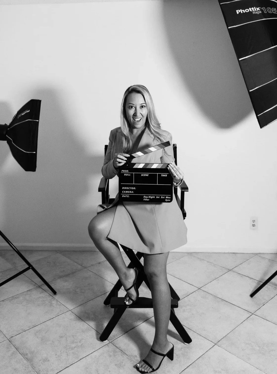 A woman sitting in a directors chair with a scene clapper smiling at the camera with professional photography lights in front of a white wall in black and white