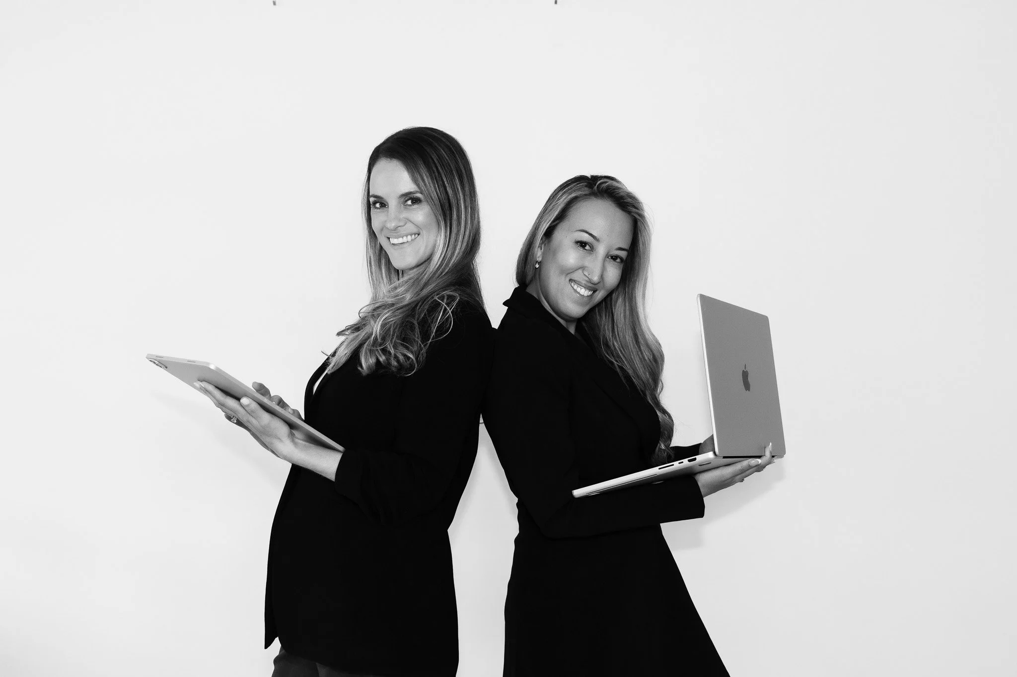 two woman wearing black dress coats hold a laptop and a tablet standing back to back