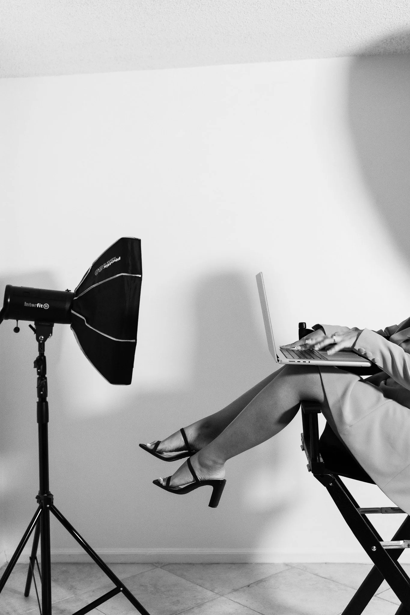 a woman types on a laptop as she sits in a chair sideways wearing heels in black and white
