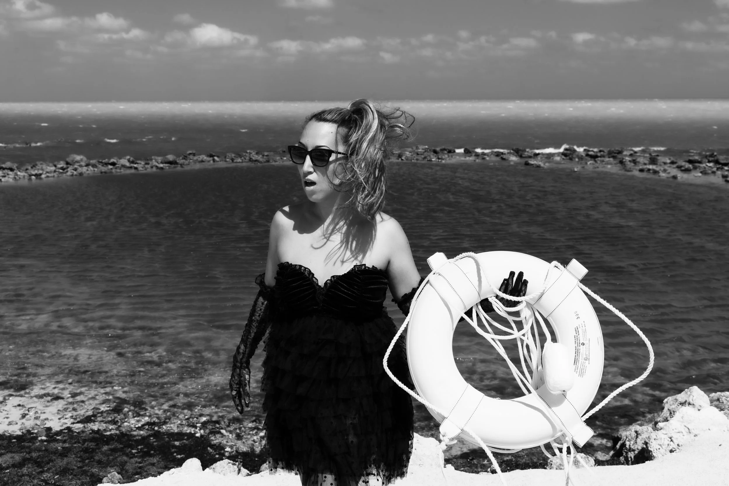 Woman in black lace dress and sunglasses holds a lifesaver floatie while standing in front of an ocean