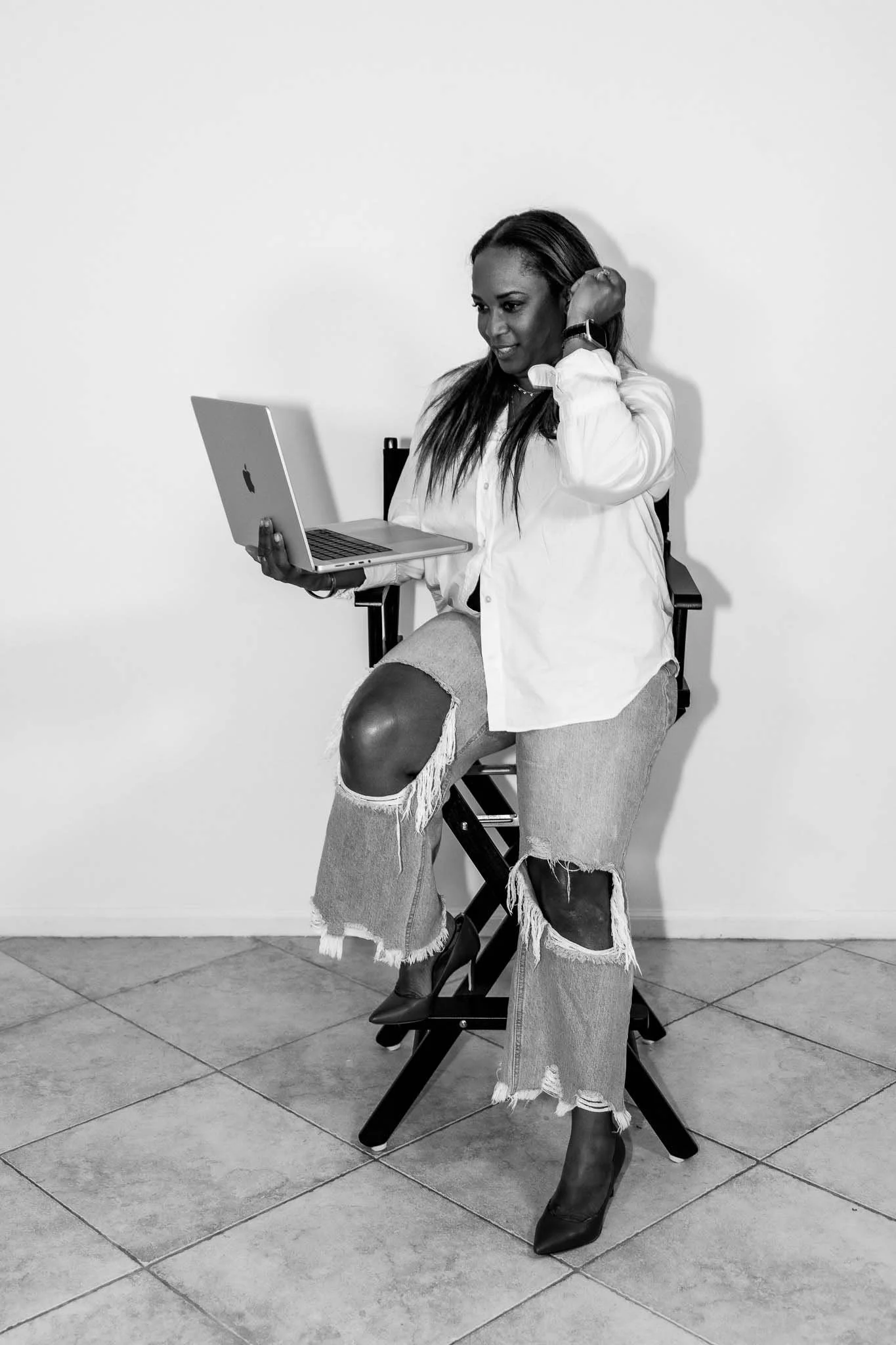 A woman sits down in a directors chair holding a laptop as she puts her hair behind her head