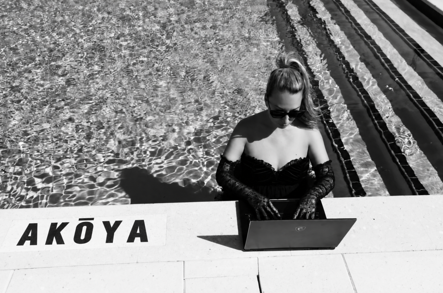 A woman in a black dress in a pool typing on a laptop with "Akoya" written on the pool deck