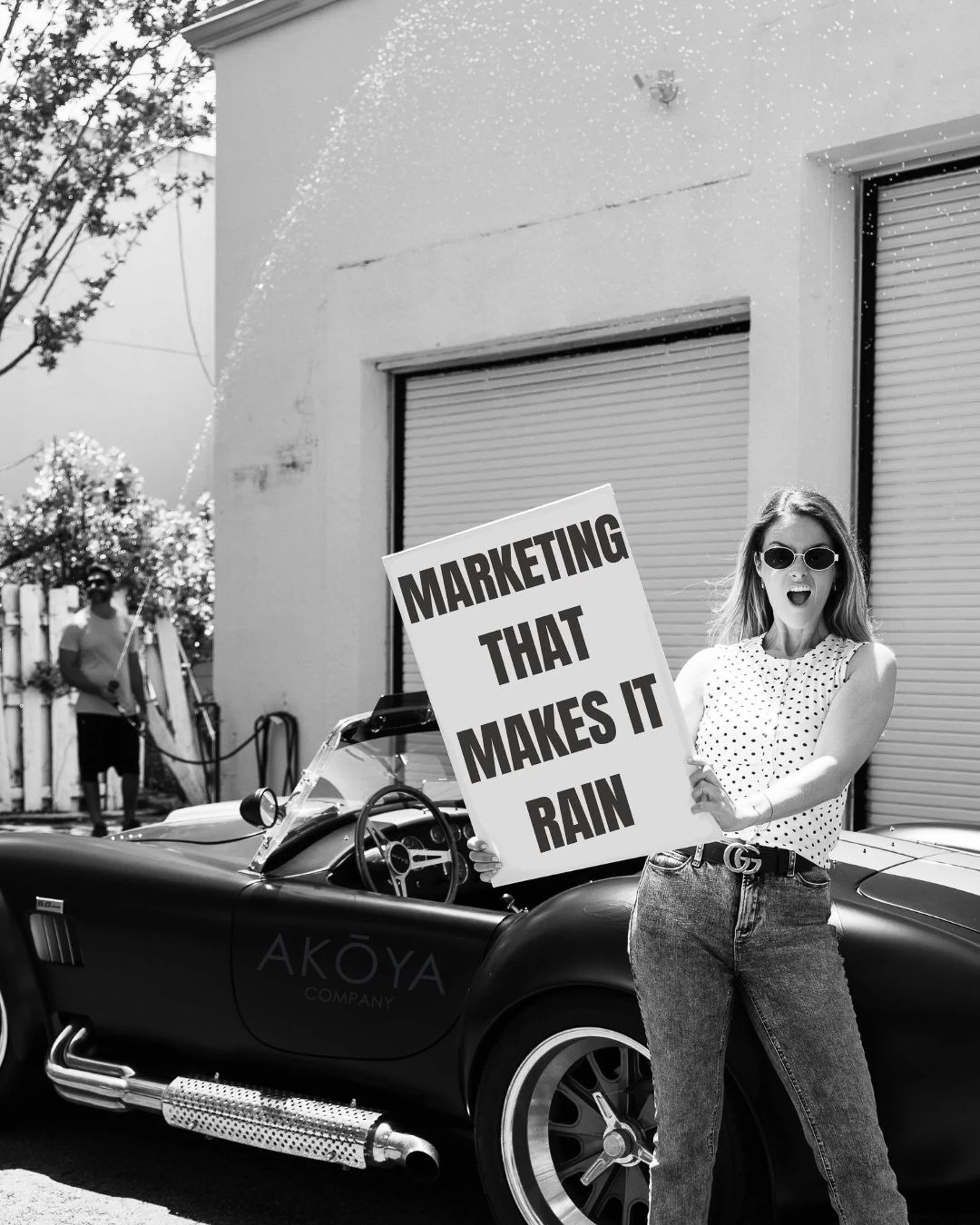 a woman outside holding a sign above her head that says marketing that makes it rain, while a man in the background hoses her down with a hose in front of a warehouse door and old school convertable car