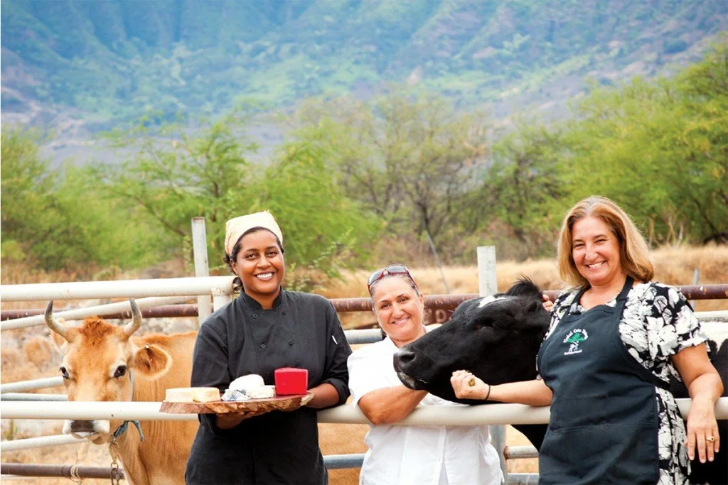 Three women standing in a rural farm area with a cow and calf, smiling and posing for a photo.
