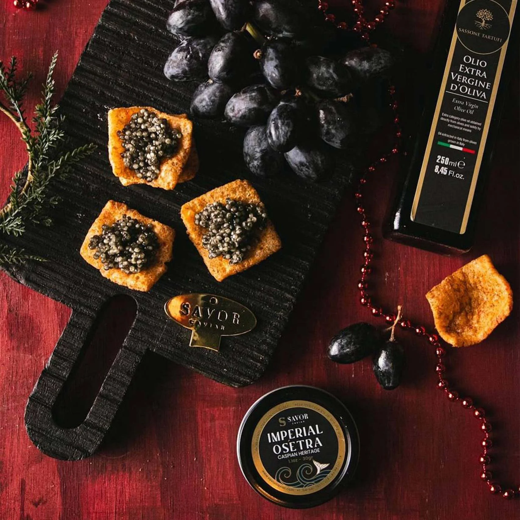 A black rectangular serving board with three square pieces of bread topped with black caviar. Next to the board, there is a bunch of black grapes, a bottle of extra virgin olive oil, and a small container labeled "Imperial Osetra" caviar. A string of red beads and some sprigs of greenery are also on the red wooden surface.