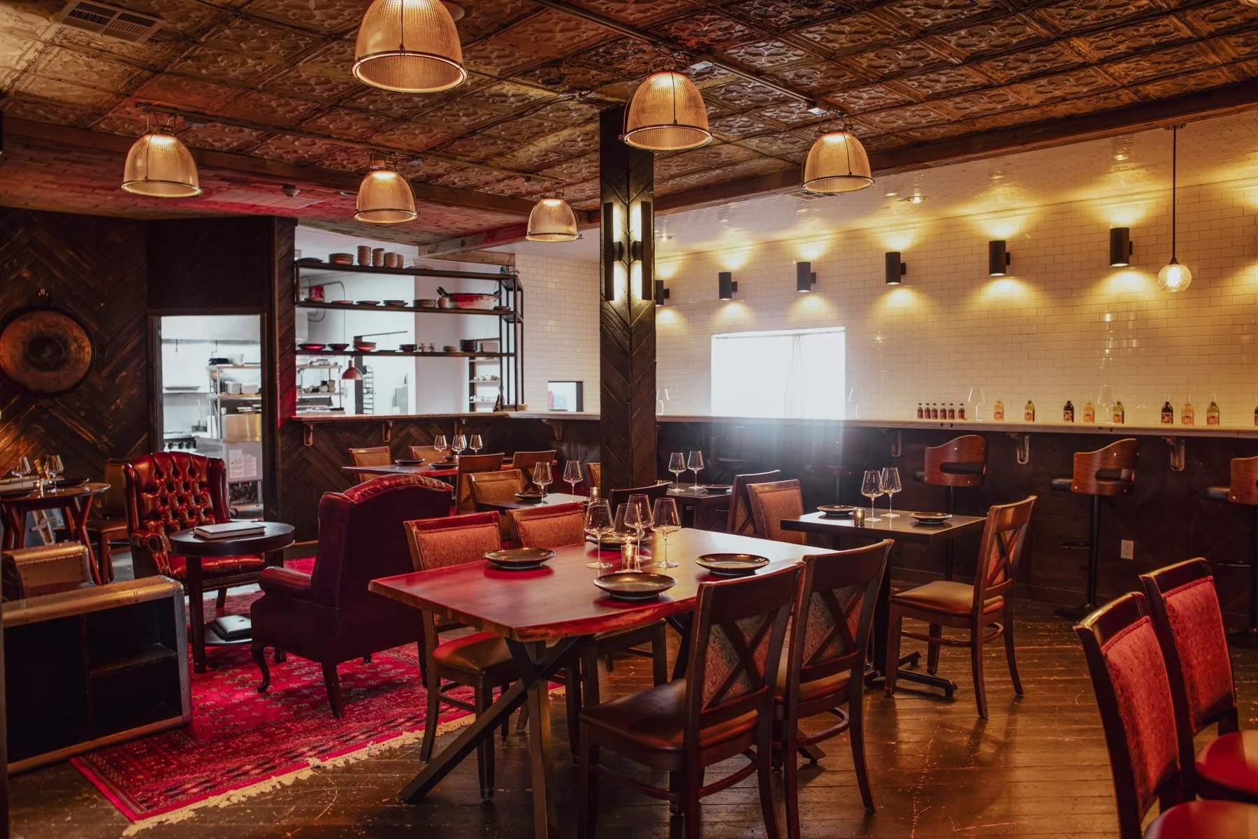 Interior of a cozy restaurant with wooden floors, red and brown leather chairs, and a mixture of vintage and modern lighting. The tables are set with black plates and wine glasses, and there is a bar area with barstools along a white-tiled wall.
