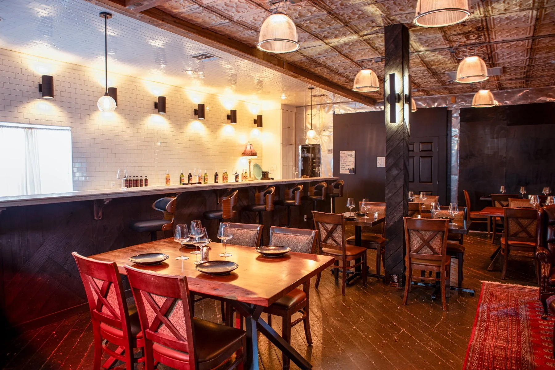 A cozy restaurant interior with wooden tables and chairs, a bar with high stools, pendant and wall lights, white subway tile walls, dark wood accents, and dim lighting for a warm atmosphere.