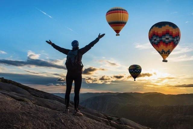 Person standing on mountain with arms raised, looking at hot air balloons