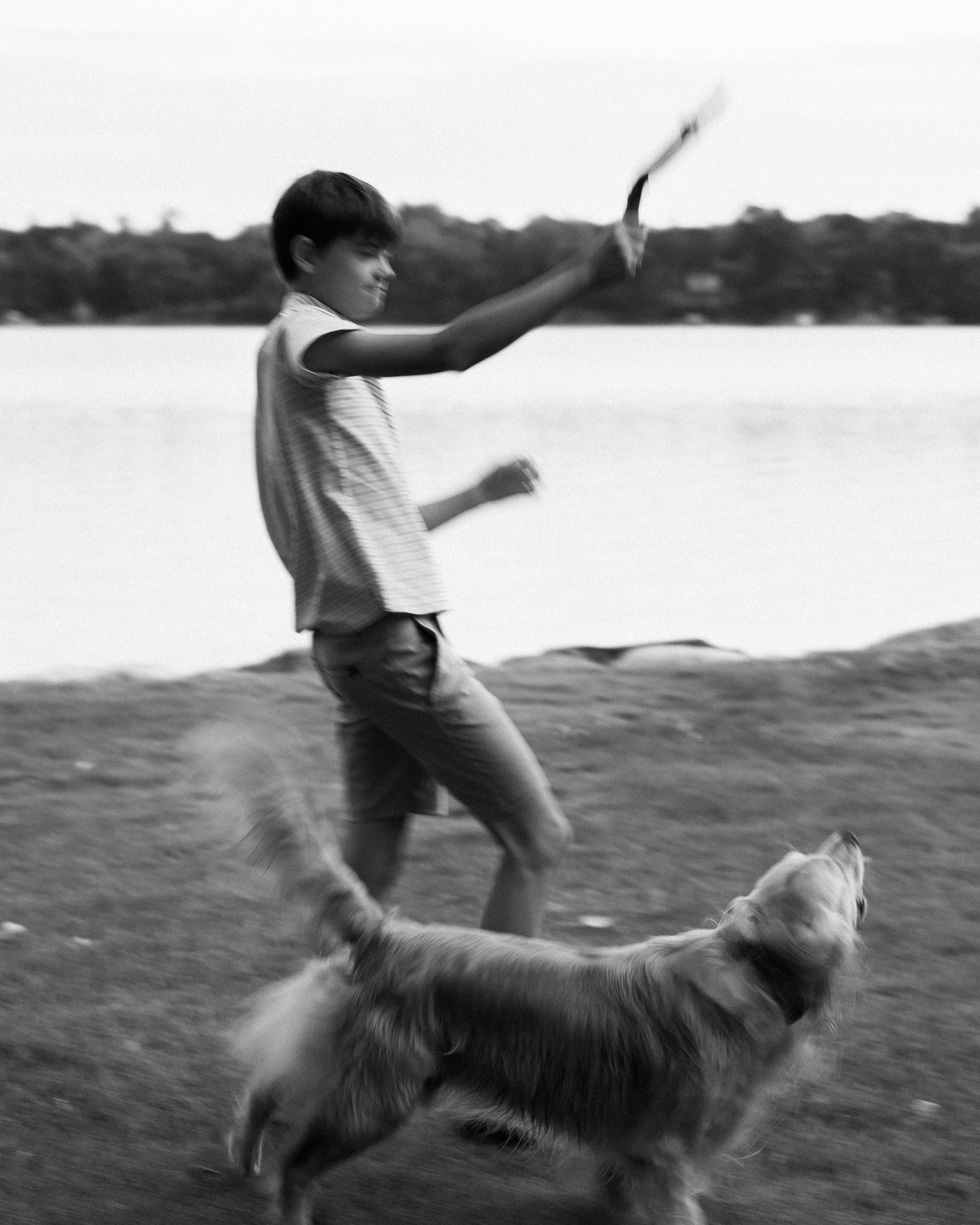 A boy running with a dog outdoors near a body of water, in black and white.