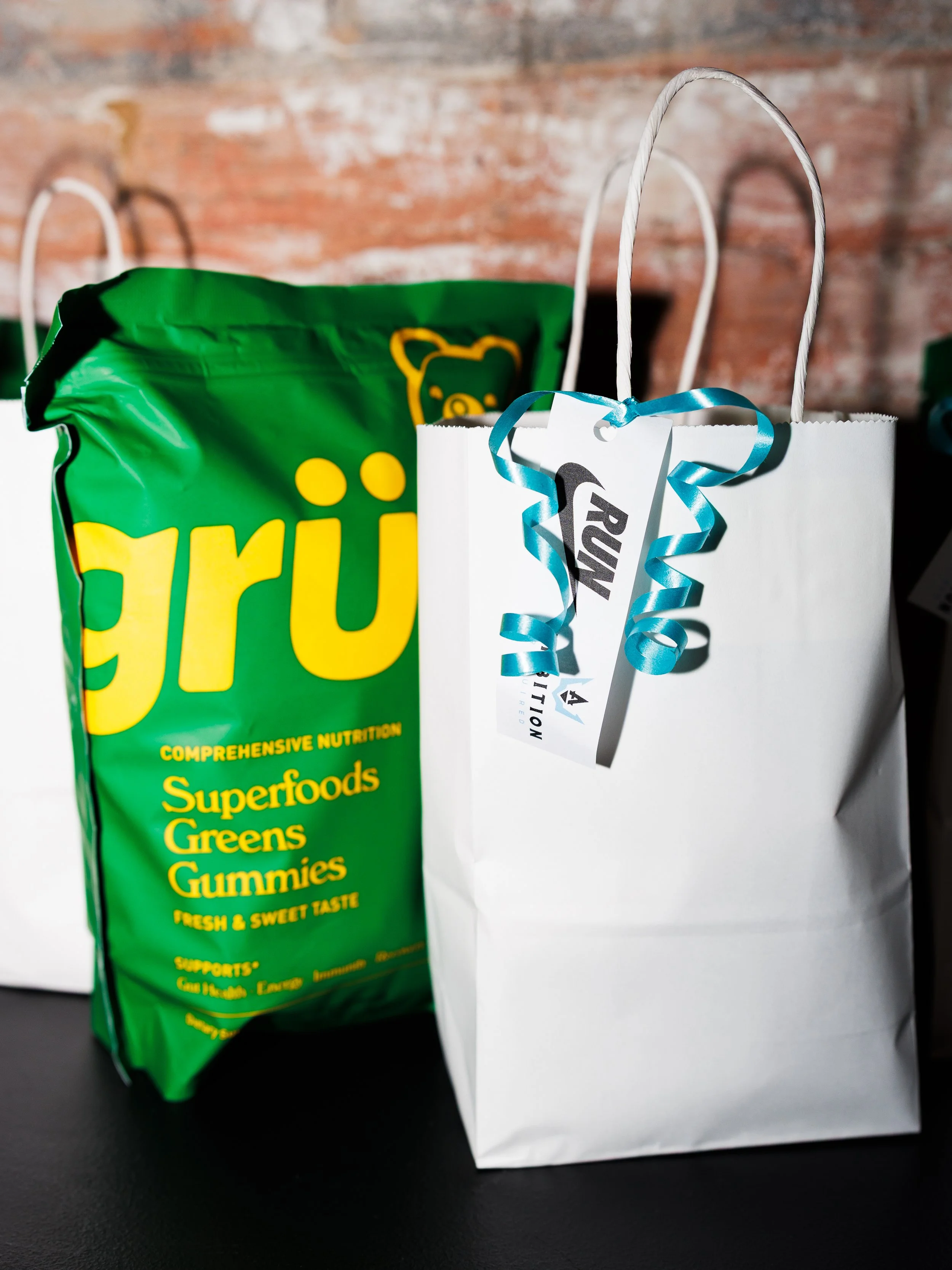 A white gift bag with a blue ribbon tag and a brown handle, standing next to a green package of supplement gummies labeled 'Superfoods Greens Gummies'.